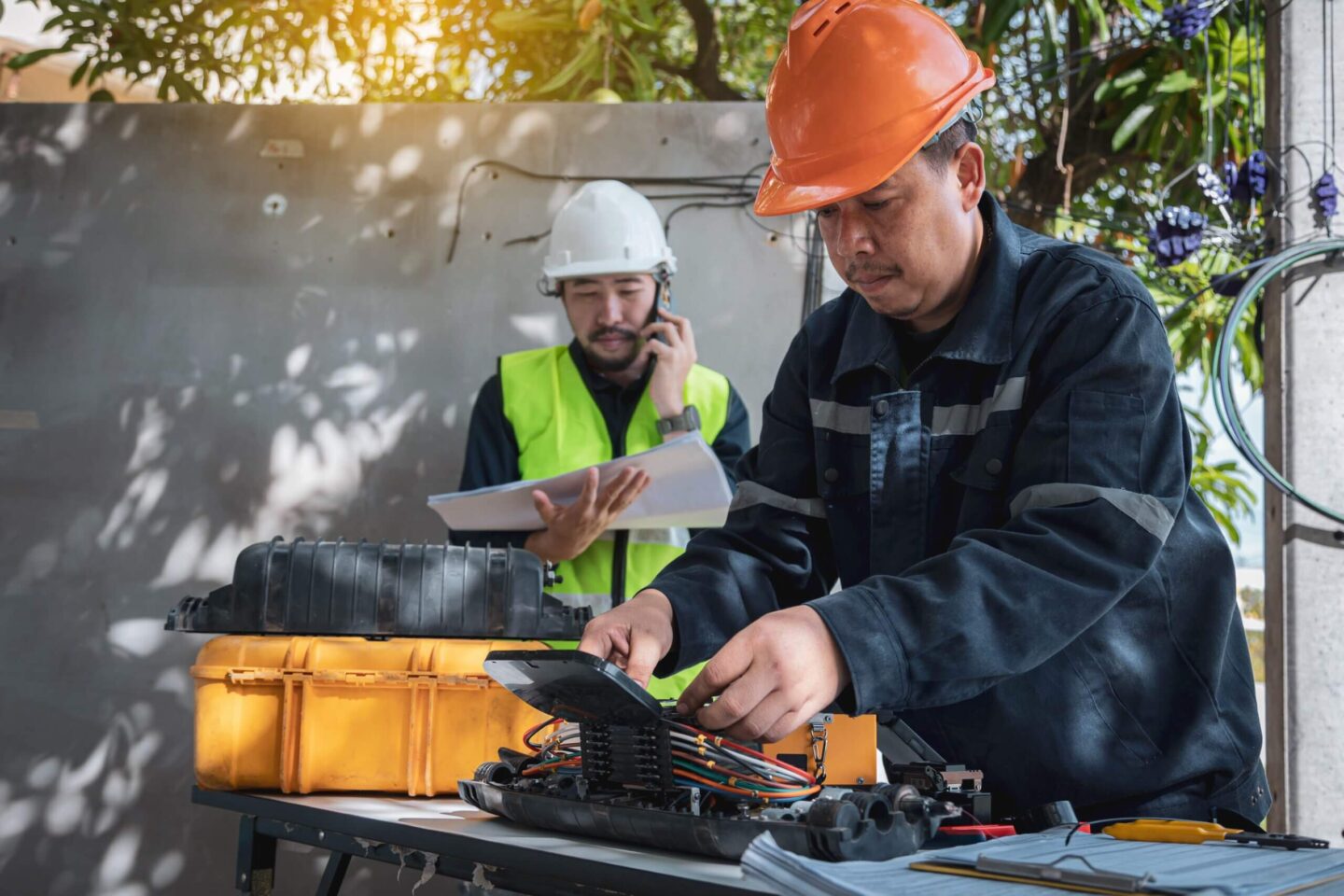 Two technicians working on fiber equipment, one inspecting cables while the other reviews documents and talks on the phone