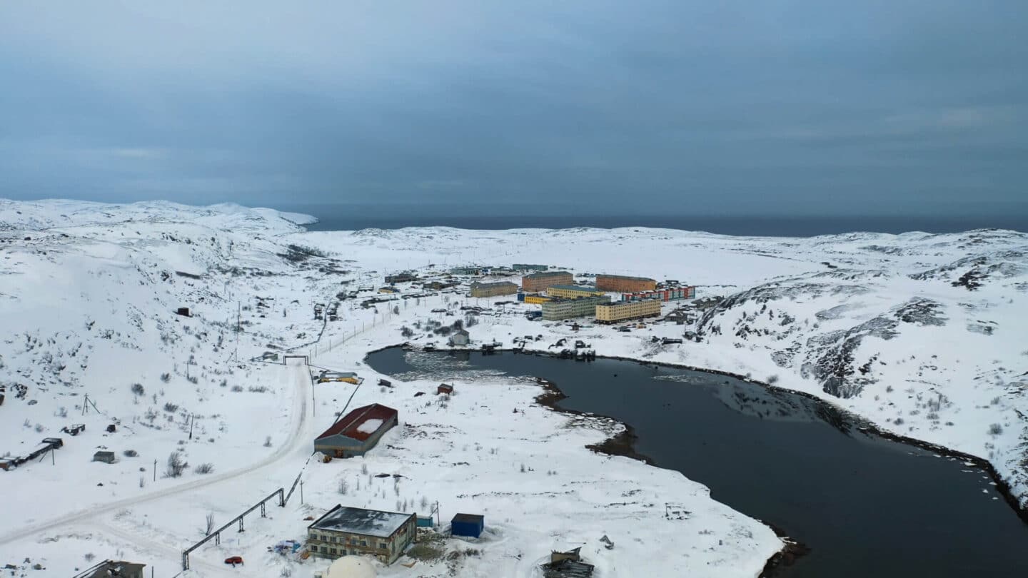 Snow-covered settlement surrounded by hills and a partially frozen lake under an overcast winter sky