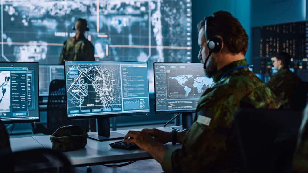 Military personnel monitoring maps and intelligence data on multiple screens in a command center