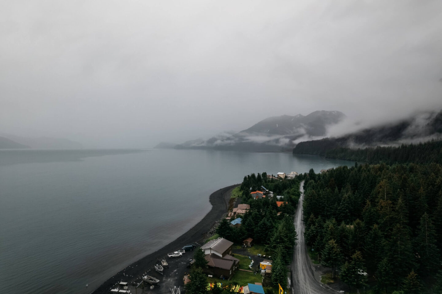 Foggy coastal landscape with a forested shoreline, mountains, and small homes along a winding road