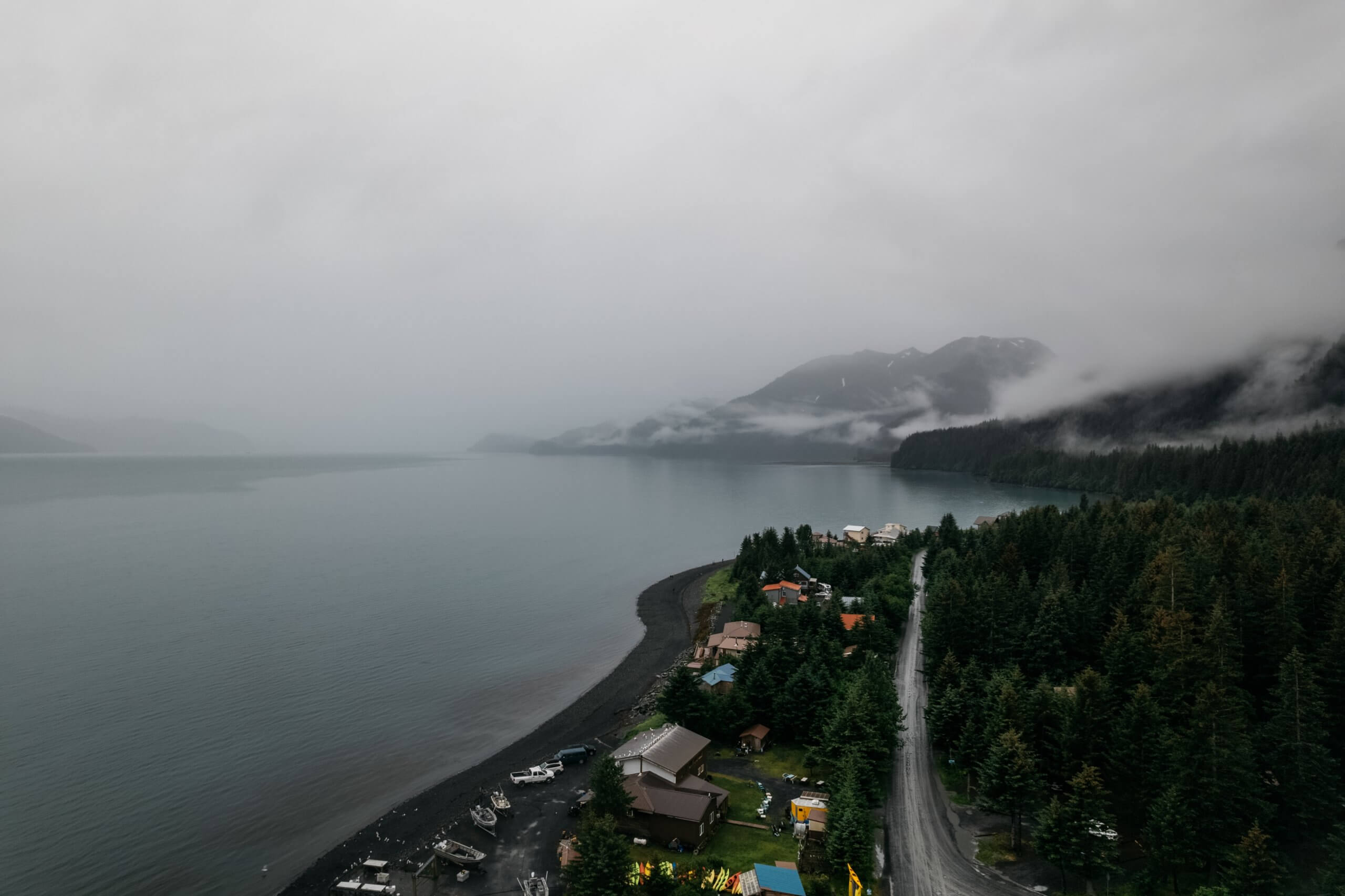 Foggy coastal landscape with a forested shoreline, mountains, and small homes along a winding road
