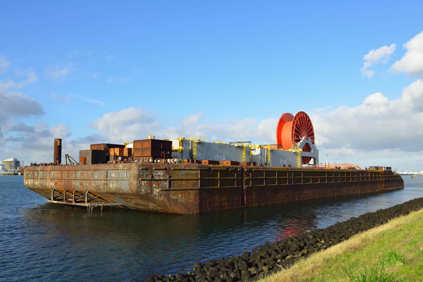 Large industrial barge with heavy equipment and a red cable reel docked along the shoreline
